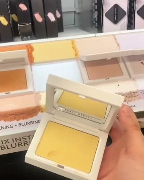 Mano sosteniendo un polvo compacto amarillo frente a un exhibidor de maquillaje